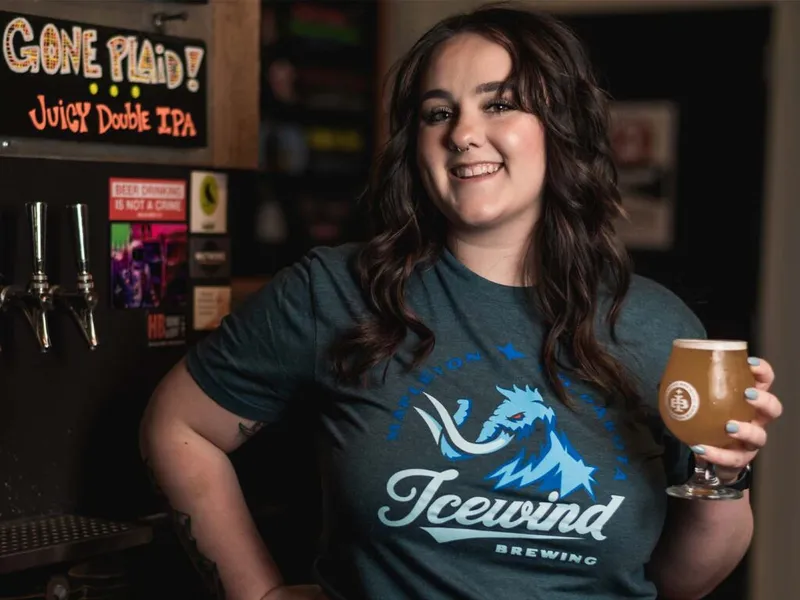 Smiling bartender in an Icewind Brewing shirt holding a pint of craft beer near tap handles