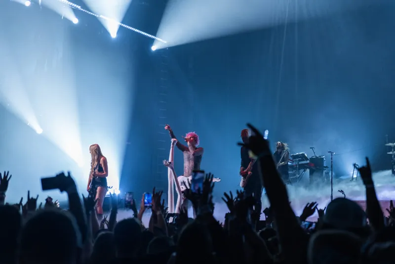 Rock performer with pink hair raises fist on stage amid blue spotlights before a crowded audience