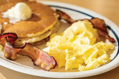 Fluffy pancakes with butter, scrambled eggs, and crispy bacon on a white plate