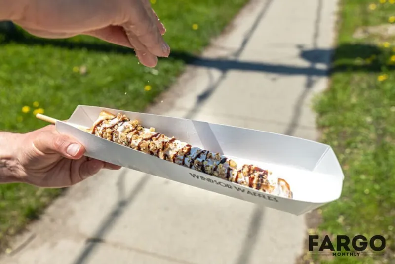 Hand holding a paper tray with a chocolate-drizzled corn dog topped with crushed nuts