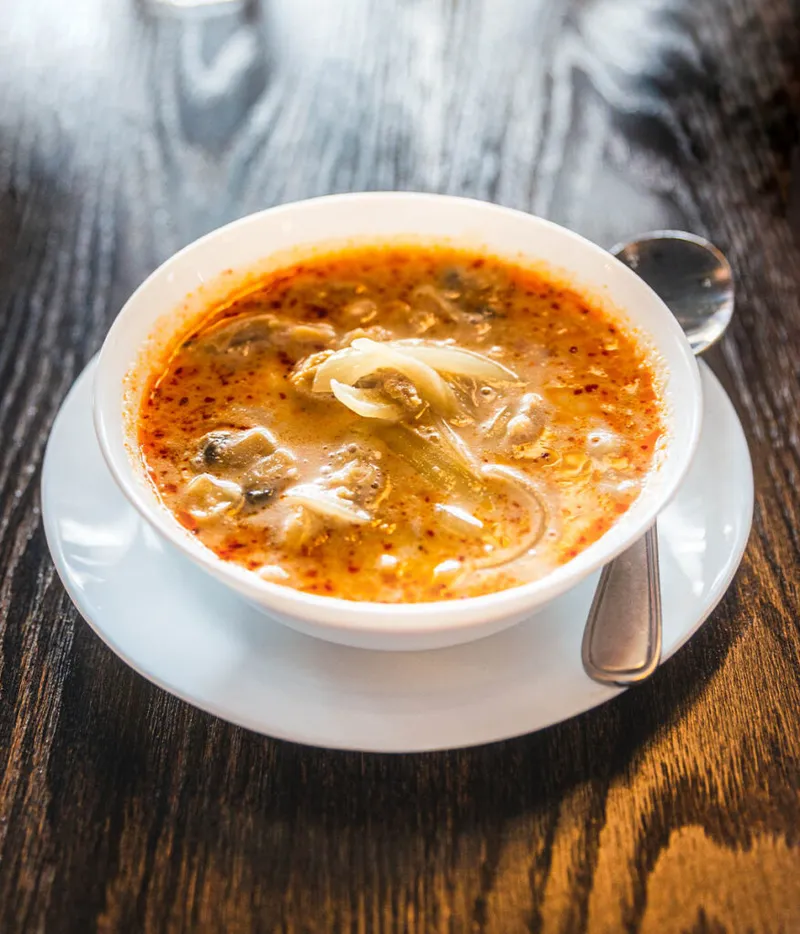 A white bowl of russet-colored brothy soup with mushrooms and onions on a saucer with a spoon, on a dark wood table