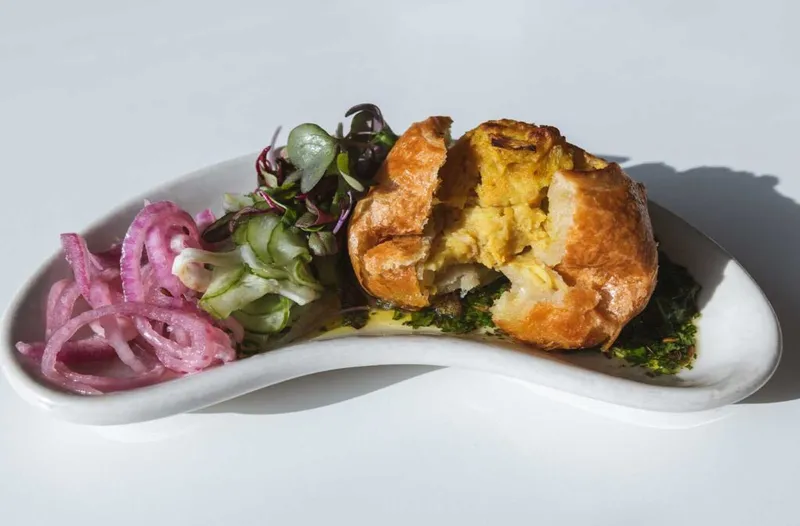 Broken golden pastry puff with herb sauce beside pickled red onions and mixed greens on a curved white dish