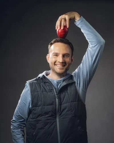 Smiling man in a blue hoodie and vest balancing a red apple on his head