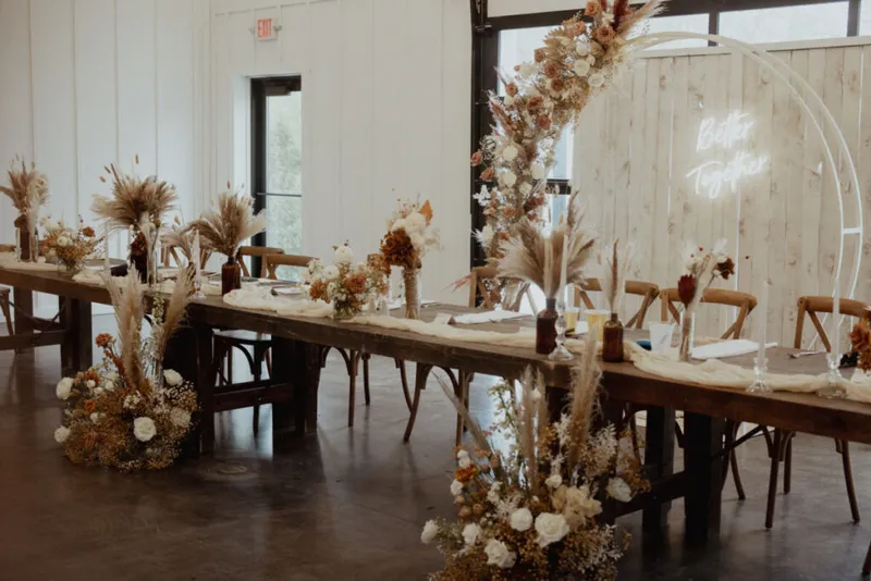 Long wooden farm table adorned with pampas grass, dried florals, and a circular neon sign in a bright white venue
