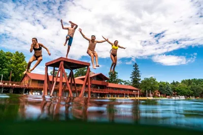 Four kids leaping off a wooden dock platform into a calm lake near a red lodge building