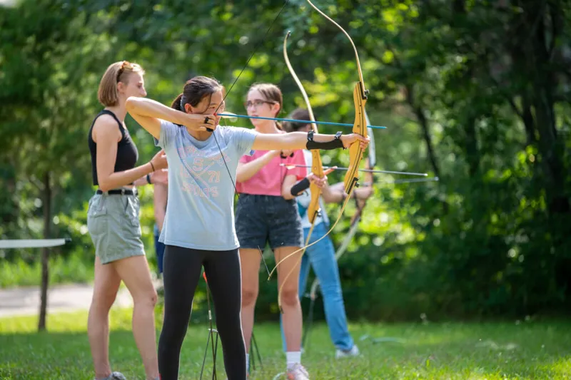 Two girls draw bows during an outdoor archery session on a sunny grass field