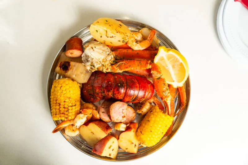 Seafood boil platter with lobster tail, crab claws, shrimp, corn, sausage, and potatoes on a metal tray