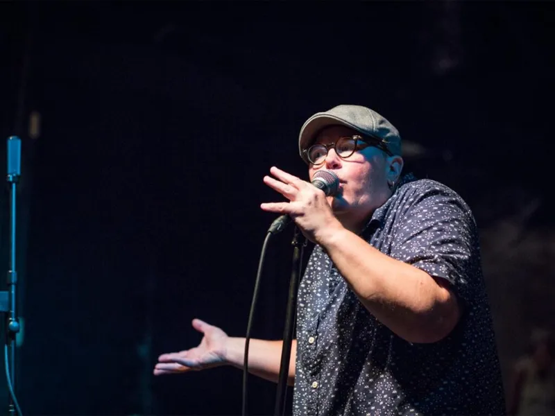 Man in flat cap and glasses singing into a mic on a darkened stage