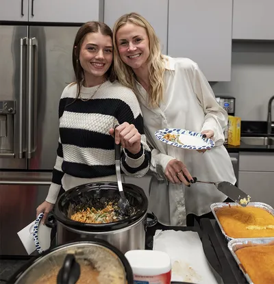 Two smiling women serving food from slow cookers and foil trays at an indoor potluck