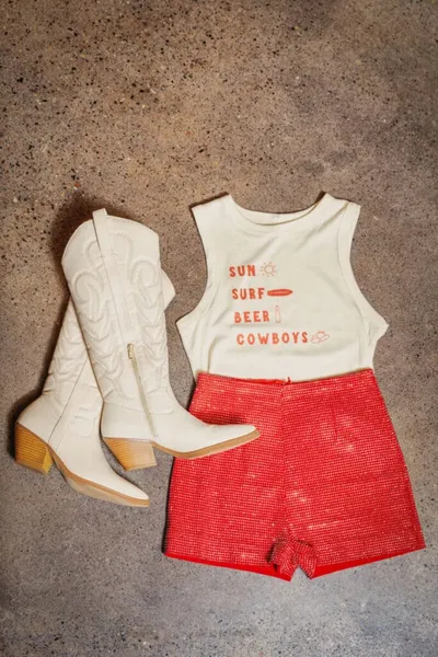 White cowboy boots beside a graphic tank top and red sparkly shorts laid flat on concrete