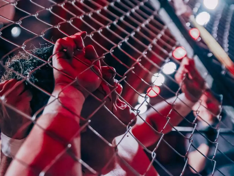 Gloved hands gripping a chain-link fence inside a pro wrestling cage under red arena lights