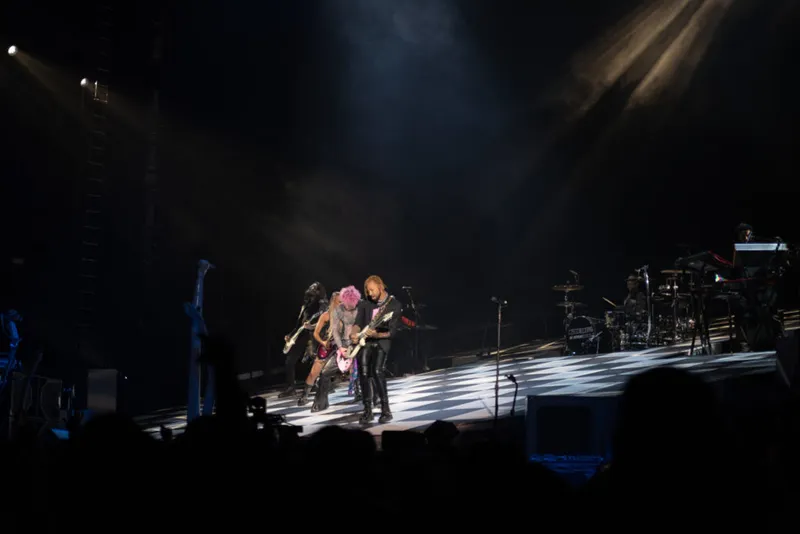 Two guitarists with pink hair perform on a darkened arena stage under dramatic spotlights