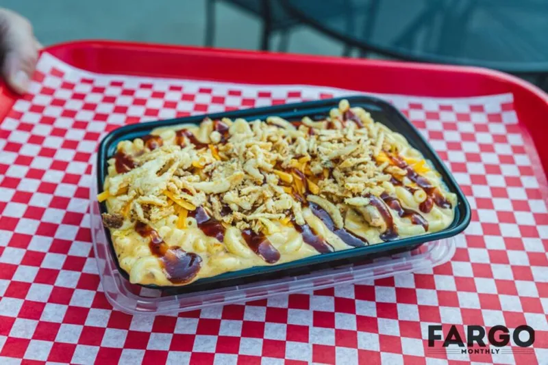 Mac and cheese topped with BBQ sauce and crispy onions in a takeout tray on a red checkered tray liner