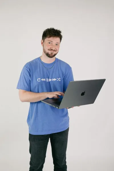 Bearded young man in blue graphic tee holding an open laptop, smiling at camera