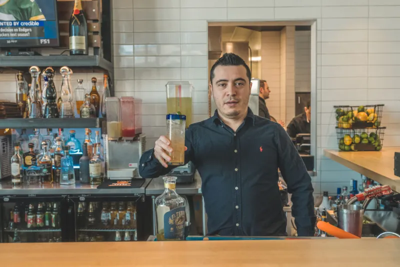 Bartender in a dark shirt holds a cocktail shaker behind a well-stocked bar