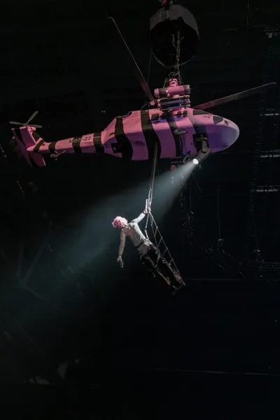 Pink-haired performer dangling from a rope beneath a pink prop helicopter on a dark stage