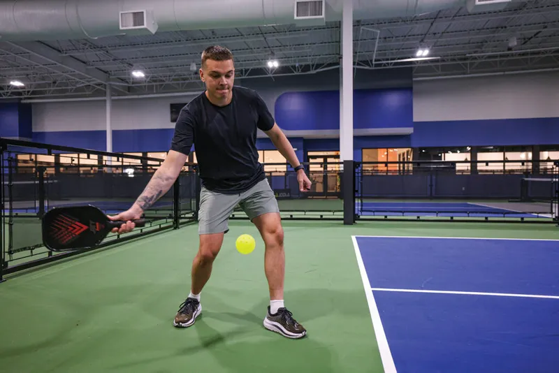 Man in black t-shirt hitting a pickleball with a red paddle on an indoor court