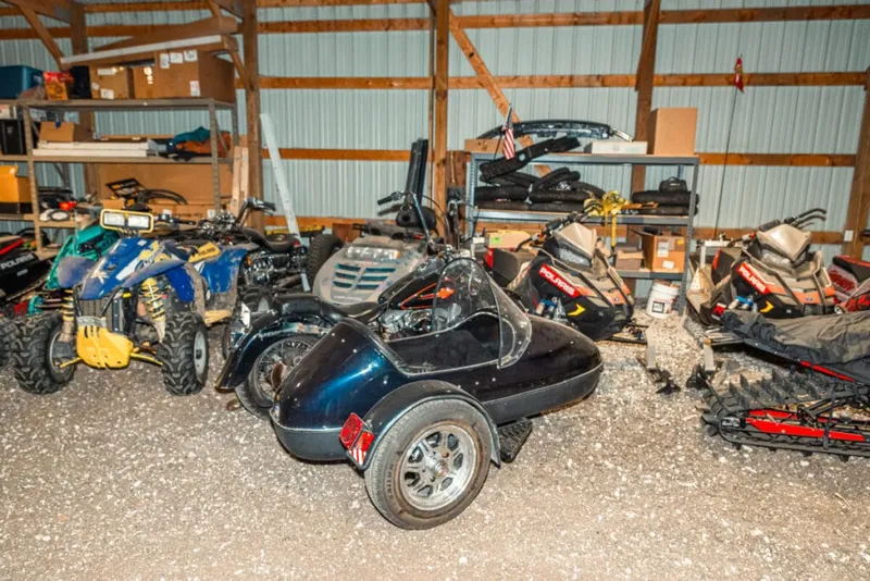 Black three-wheeled kit car parked among ATVs and snowmobiles inside a metal storage barn