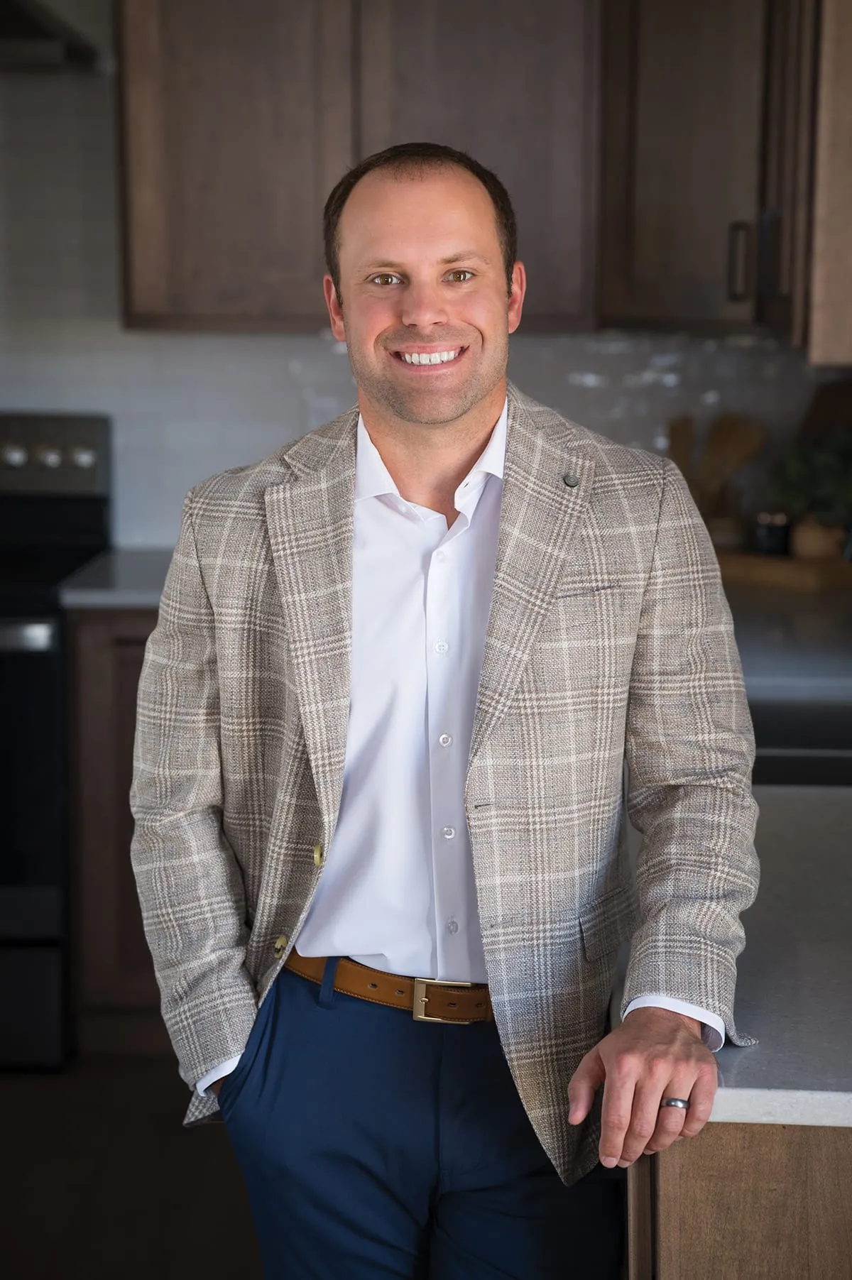 Man in plaid blazer and navy trousers leaning against a kitchen counter, smiling