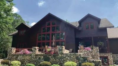 Large dark-wood lodge with red-trimmed windows and a stone retaining wall lined with pink flowers