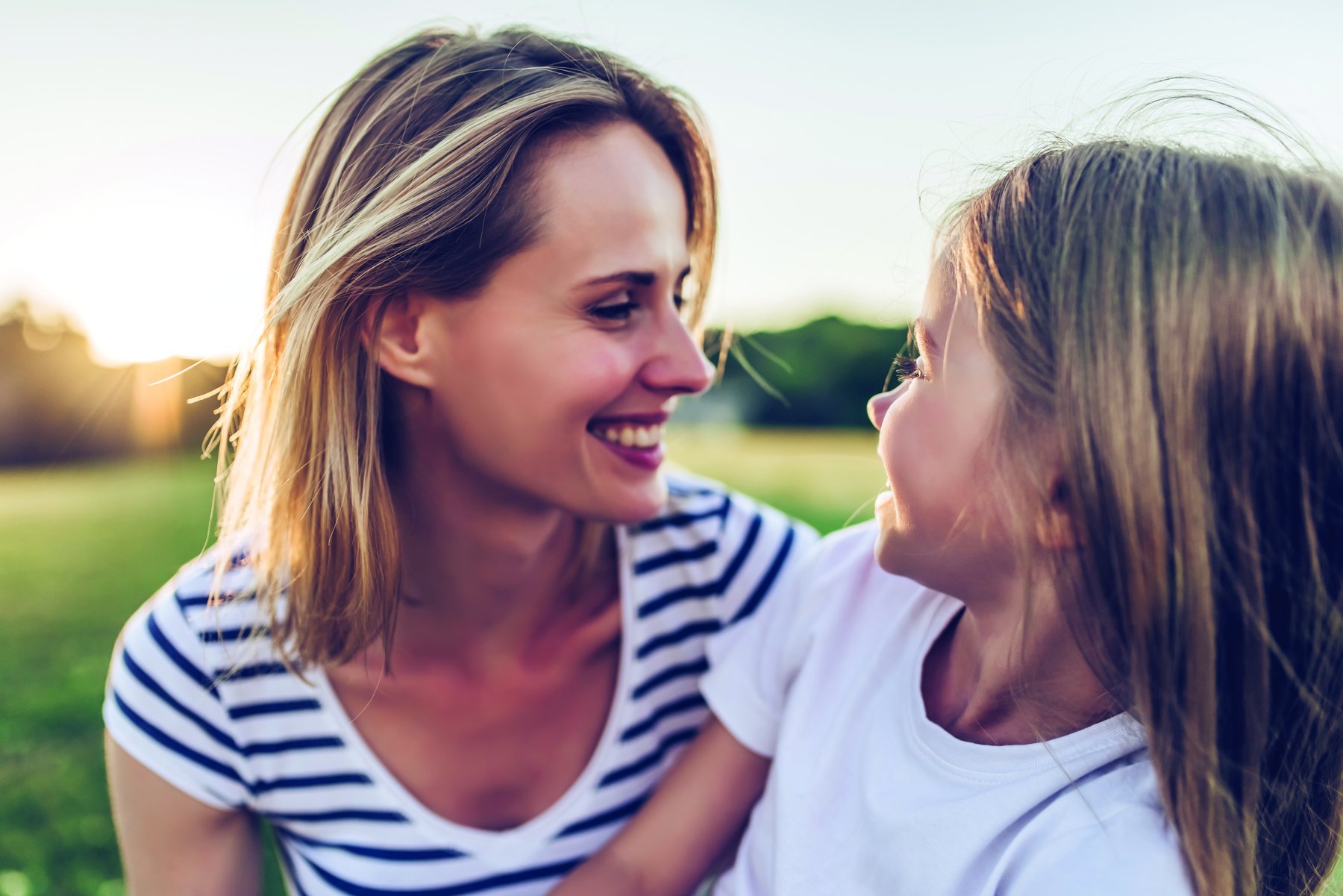 A smiling mom and her daughter look at each other outdoors at sunset