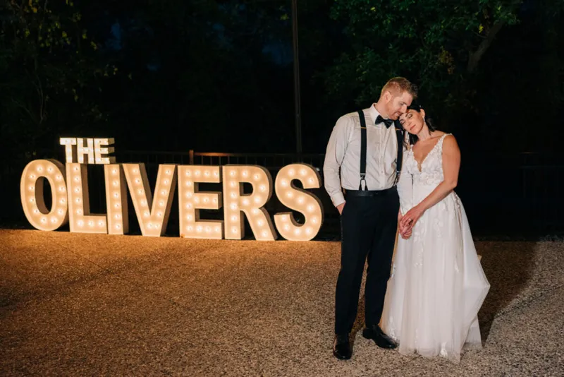 Bride and groom embrace at night beside large illuminated marquee letters reading 'The Olivers