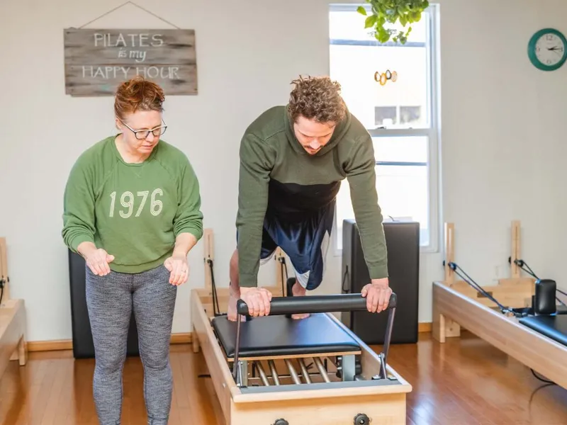 Pilates instructor in green sweatshirt observes man leaning over a reformer in a bright studio