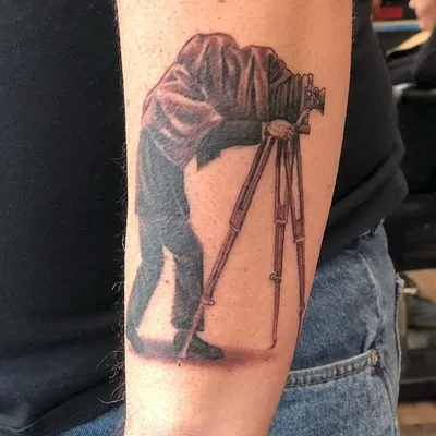 Black and grey forearm tattoo of a vintage photographer hunched over a tripod camera