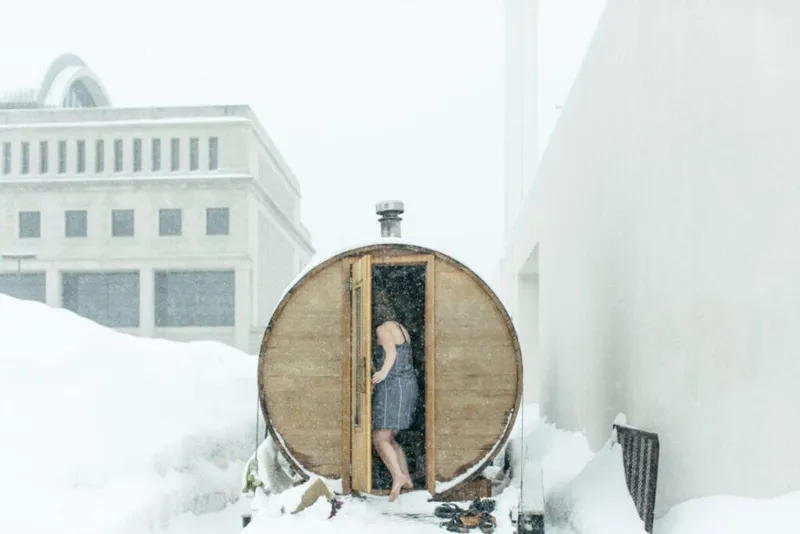 Woman in a swimsuit stepping into a wooden barrel sauna surrounded by deep snow during a blizzard