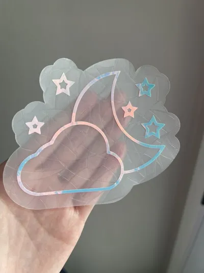 Hand holding a translucent holographic sticker with a crescent moon, cloud, and stars