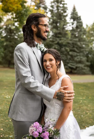 Bride in white lace gown holds purple flowers as groom in gray suit embraces her outdoors