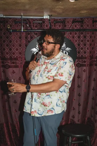 Curly-haired man in a floral shirt speaks into a mic while holding a drink on a small stage