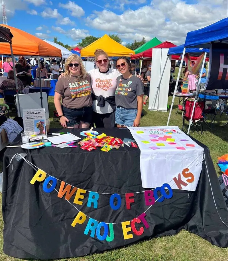 Three women smiling behind a Power of Books Project booth at an outdoor festival