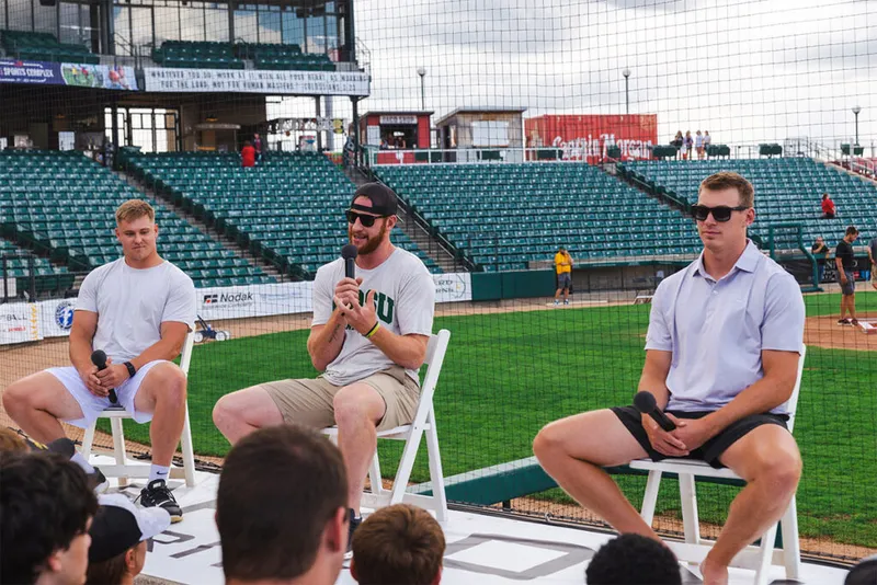Three young men seated in chairs on a baseball field holding microphones for a panel discussion