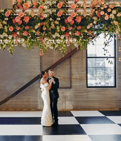 Couple sharing first dance beneath a lush floral chandelier on a black-and-white checkered floor