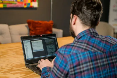 Bearded man in plaid shirt viewed from behind, typing on a laptop showing code