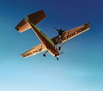 Skydiver standing atop a small plane in flight against a clear blue sky