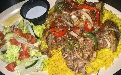 Grilled meat over yellow rice with sautéed peppers and onions, served alongside a garden salad and white dipping sauce