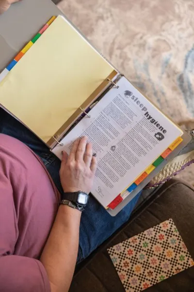 Person reading a tabbed binder open to a sleep hygiene page, seated on a couch