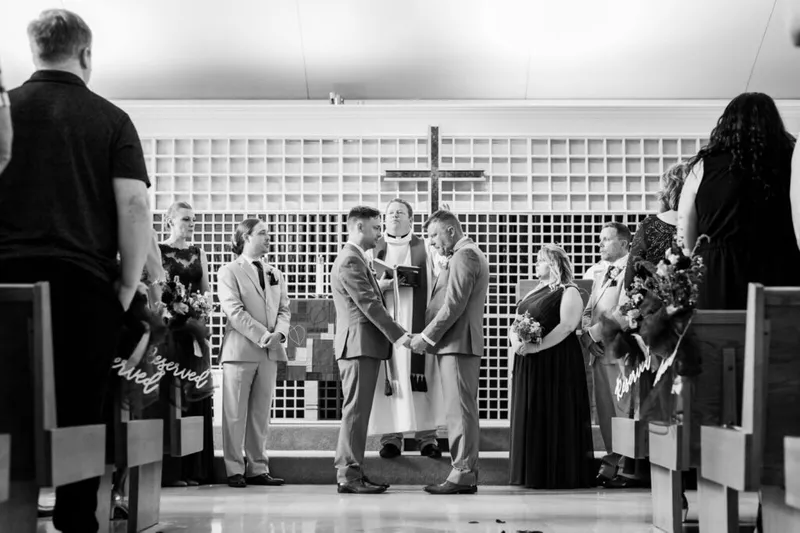 Two grooms hold hands at the altar before an officiant in a church ceremony