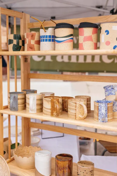 Handmade ceramic mugs and painted travel cups displayed on a wooden shelving unit at a market stall