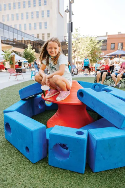 Young girl perched on a red play structure surrounded by blue foam blocks on artificial turf in an urban plaza