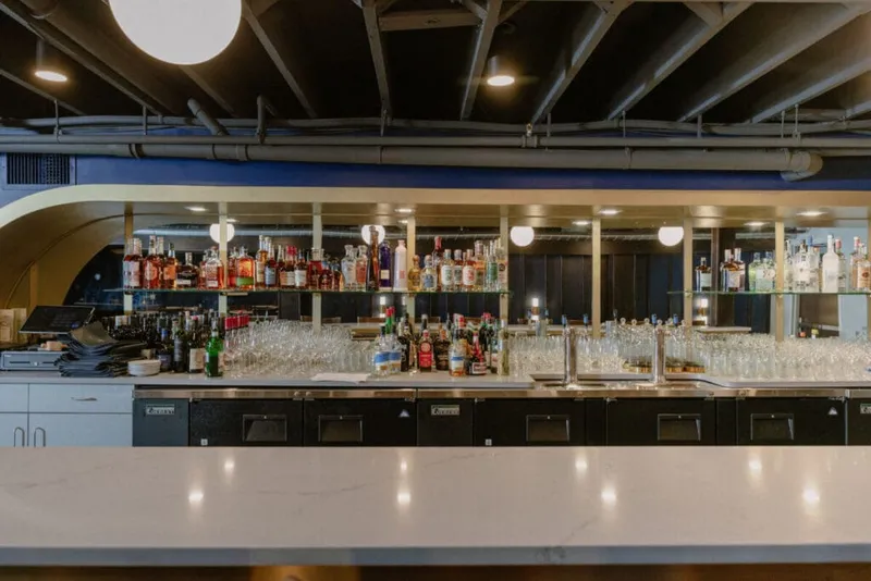 Marble bar counter with shelves of liquor bottles and glassware under an exposed ceiling