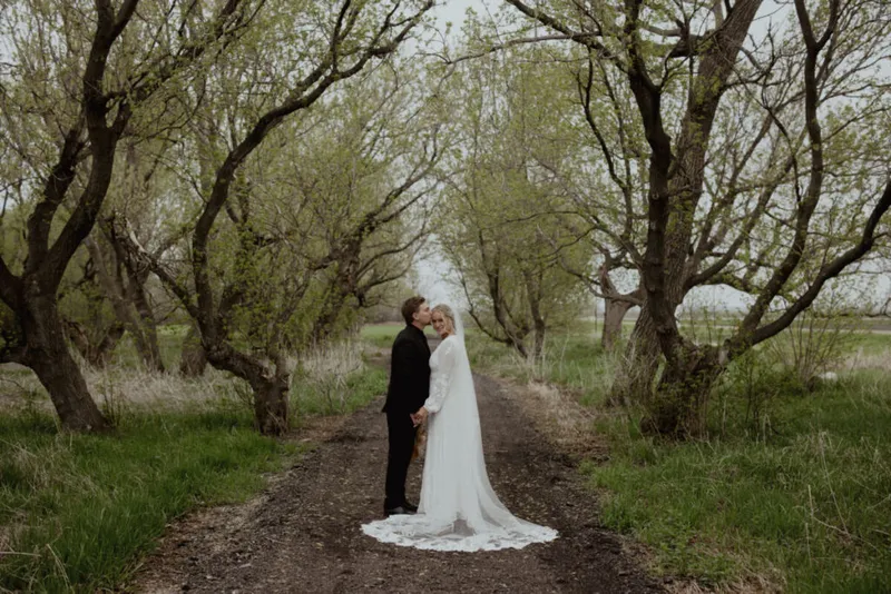 Couple sharing a kiss on a tree-lined dirt path, bride in long white gown with veil