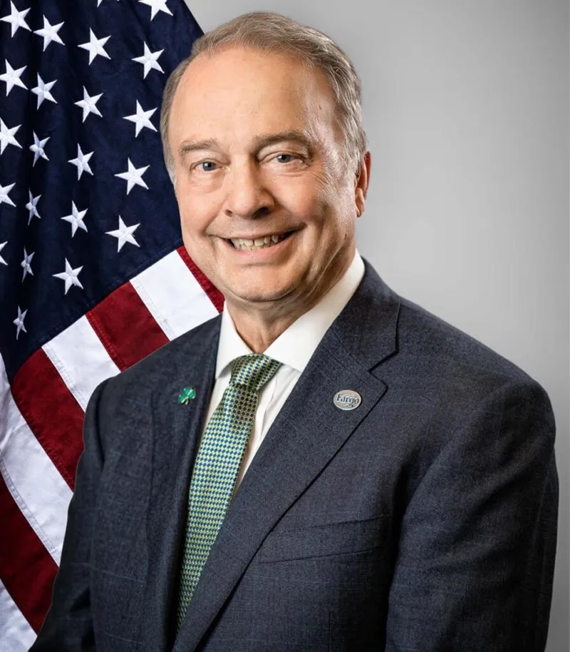 Smiling man in gray suit and green tie poses before an American flag with a Fargo lapel pin