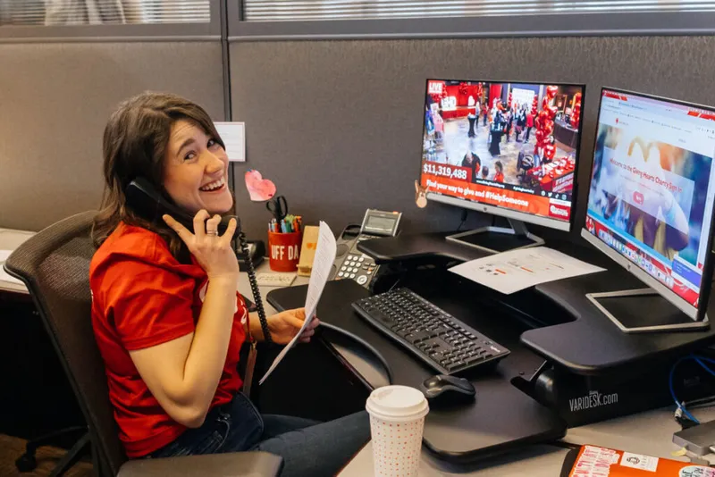 Smiling woman in red shirt talking on phone at cubicle desk with charity telethon on dual monitors