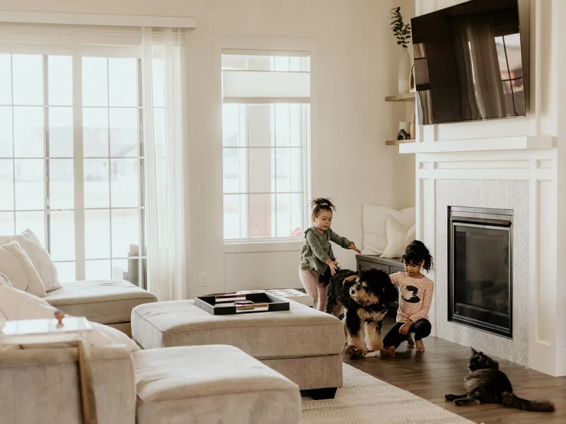 Two young girls petting a large dog near a white fireplace in a bright living room, with a cat resting nearby
