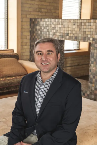 Smiling man in dark blazer and checked shirt seated in a warmly decorated interior