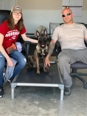 Woman in red shirt and man in sunglasses sit on either side of a Belgian Malinois on a cot