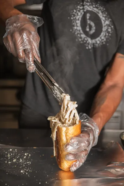 Gloved hands using tongs to pile steaming shredded meat onto a hoagie roll